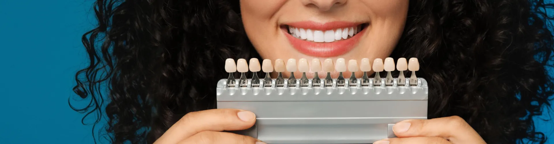 Smiling woman holding a dental shade guide in front of her bright white teeth to compare tooth color for cosmetic veneer treatment.