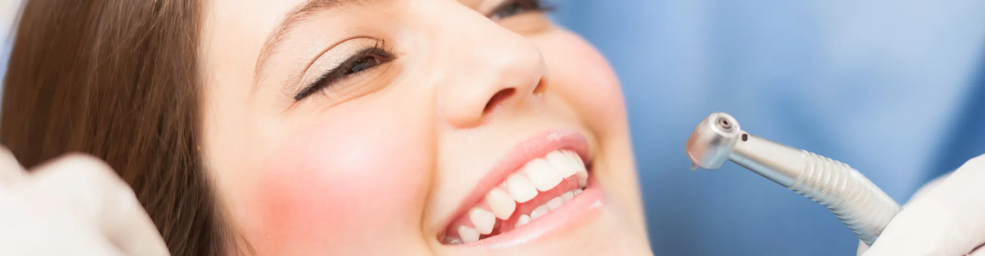 Gloved hands holding dental tools around smiling woman's mouth.